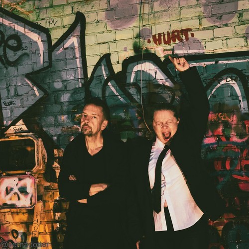 Rymdverket – Numb single cover: the duo in black in front of a graffiti-covered brick wall, one with arms folded, the other in a white shirt and tie with a fist raised and mouth wide open