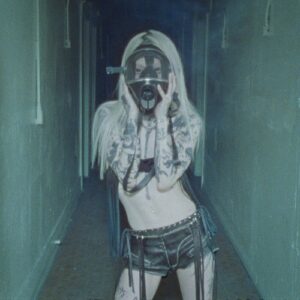 yeule – Hystérique single cover: a washed-out blue photo of yeule in a narrow corridor, platinum hair, tattooed arms raised to a gas mask, wearing low-slung fringed shorts